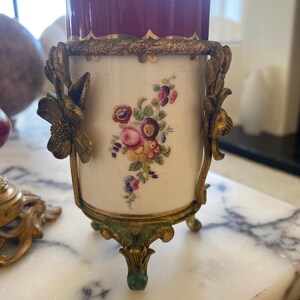 May include: An antique decorative vase with a burgundy interior and a white exterior adorned with a floral design. The vase is held by a gold-colored metal frame with floral accents and rests on three ornate feet.