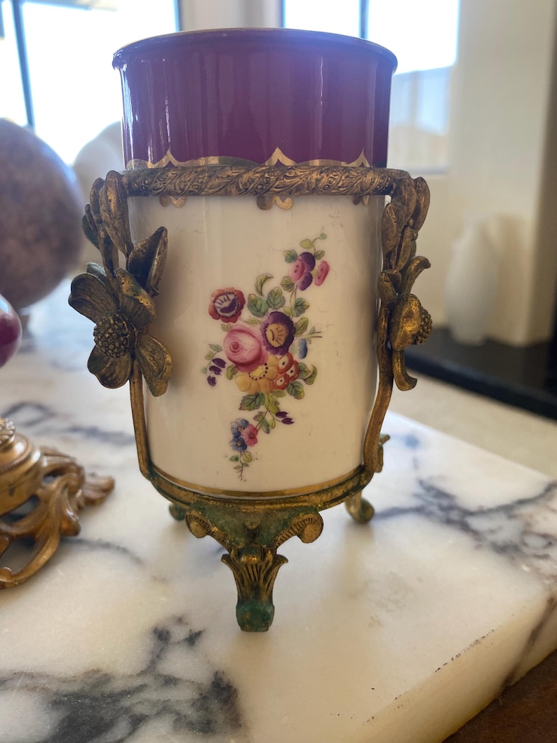 May include: An antique decorative vase with a burgundy interior and a white exterior adorned with a floral design. The vase is held by a gold-colored metal frame with floral accents and rests on three ornate feet.