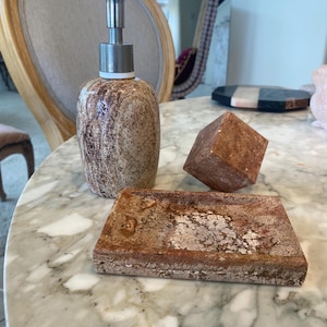 May include: A bathroom set featuring a brown and beige speckled soap dispenser with a silver pump, a rectangular soap dish, and a geometric cube. The items are displayed on a marble surface.