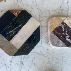 May include: Two decorative coasters. One is an octagon shape, and the other is square with rounded corners. Both are made of marble and feature a geometric design with black, white, green, brown, and beige tones. The coasters are on a marble surface.