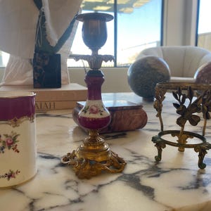 May include: An antique collection of decorative items on a marble surface. Includes a white and burgundy floral ceramic cup, a brass and porcelain candlestick, and a small brass stand with floral accents. The items are vintage.