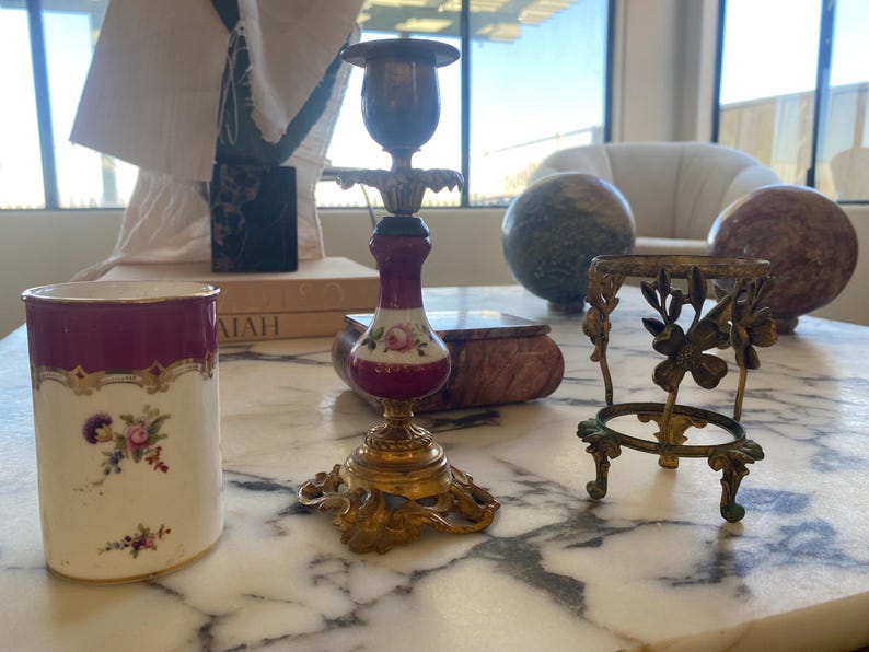 May include: An antique collection of decorative items on a marble surface. Includes a white and burgundy floral ceramic cup, a brass and porcelain candlestick, and a small brass stand with floral accents. The items are vintage.