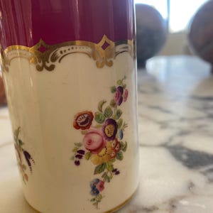 May include: A cylindrical, antique-style mug with a deep burgundy top and a cream-colored body. The mug features gold filigree accents and a floral bouquet design in shades of pink, purple, and yellow. The base has a gold trim.