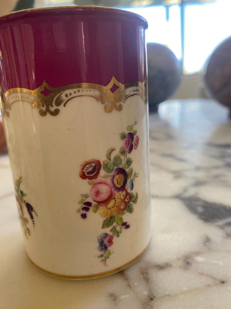 May include: A cylindrical, antique-style mug with a deep burgundy top and a cream-colored body. The mug features gold filigree accents and a floral bouquet design in shades of pink, purple, and yellow. The base has a gold trim.