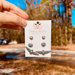 May include: A set of three silver earrings with turquoise stones. The earrings feature a starburst design, a floral design, and a bird design. The earrings are displayed on a white card with the text "The Rowdy Rancher Western Boutique".