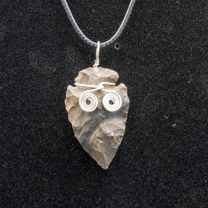 Può includere: Una collana con un ciondolo a forma di punta di freccia marrone con accenti di filo d'argento. La punta di freccia è attaccata a un cordoncino nero.