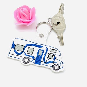 Könnte beinhalten: Ein weißer Wohnmobil-Schlüsselanhänger mit blauen Details und einem silbernen Schlüsselring. Das Wohnmobil-Design hat blaue Akzente und graue Fenster. Eine rosa Rose und zwei Schlüssel sind ebenfalls vorhanden.