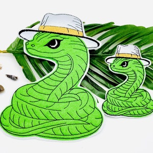 Könnte beinhalten: Zwei grüne Schlangenaufnäher, einer groß und einer klein, beide tragen weiße Fedora-Hüte mit gelben Bändern. Die Schlangen haben schwarze Augen und sind mit schwarzen Konturen bestickt. Die Aufnäher befinden sich auf weißem Hintergrund mit einem grünen Blatt.