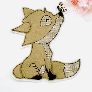 Könnte beinhalten: Ein fuchsförmiges Applikation aus hellbraunem Stoff mit einer glitzernden, goldenen Schnauze und einem Schwanz, der einen bunten Schmetterling betrachtet. Der Fuchs sitzt, mit schwarzen Pfoten und einem detaillierten Design. Rosa Blüten im Hintergrund.