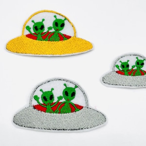 Könnte beinhalten: Drei gestickte Aufnäher mit UFOs und grünen Außerirdischen. Die UFOs sind in Gelb, Grau und Silber gehalten, die Außerirdischen in Grün und Rot. Die Aufnäher befinden sich auf weißem Hintergrund.
