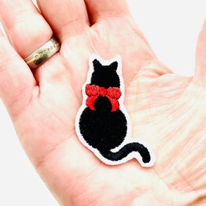 Könnte beinhalten: Gestickter schwarzer Katzenaufnäher mit einer leuchtend roten Schleife. Die Katze sitzt mit eingerolltem Schwanz. Der Aufnäher hat einen weißen Rand. Geeignet für Kleidung, Taschen oder andere Stoffartikel.