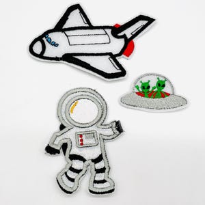Könnte beinhalten: Gestickte Weltraum-Patches mit einem weißen und schwarzen Shuttle, einem Astronauten im Raumanzug und einer grauen UFO mit grünen Außerirdischen. Die Patches haben detaillierte Nähte und sind vor einem weißen Hintergrund platziert.