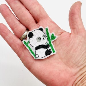 Könnte beinhalten: Gestickter Panda-Bären-Aufnäher in Schwarz, Weiß und Silber, mit grünen Bambusstängeln. Der Aufnäher ist klein und detailliert und zeigt ein niedliches Panda-Design. Der Aufnäher ist wahrscheinlich für Kleidung oder Accessoires.