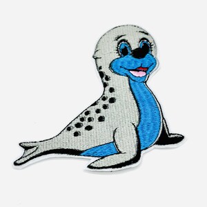 Könnte beinhalten: Gestickter Aufnäher mit einem Cartoon-Robbenmotiv. Die Robbe hat einen grauen Kopf und Körper mit schwarzen Punkten, einen blauen Bauch und ein lächelndes Gesicht mit rosa Zunge. Der Aufnäher ist zum Aufbügeln oder Aufnähen auf Stoff gedacht.
