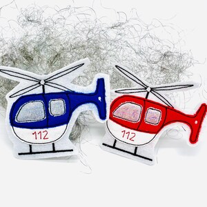 Könnte beinhalten: Zwei Filz-Hubschrauber-Ornamente, einer blau und einer rot, mit weißen Akzenten. Jeder hat ein silbernes Glitzerfenster und die Nummer "112" auf der Unterseite aufgedruckt. Die Hubschrauber haben schwarze Rotorblätter und eine weiße Basis.