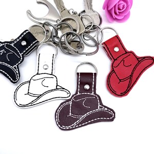 Könnte beinhalten: Eine Sammlung von vier Schlüsselanhängern in Form von Cowboyhüten in Schwarz, Weiß, Dunkelbraun und Rot, jeweils mit einem Metallring und einem silberfarbenen Druckknopf. Der schwarze Anhänger ist mit dem Buchstaben K bestickt. Eine künstliche rosa Rose.