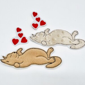 Könnte beinhalten: Vier Stoffapplikationen auf weißem Hintergrund. Zwei sind tan-farbene Nashornformen, eine mit einem gepunkteten Muster. Zwei weiße Formen zeigen rote Herzmotive. Alle haben schwarze, genähte Umrisse.