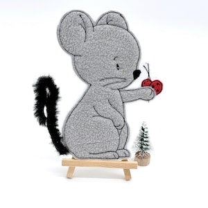 Könnte beinhalten: Eine graue Filz-Maus-Figur, die zwei rote Kirschen hält. Die Maus hat einen schwarzen Schwanz und steht auf einer kleinen Holzstaffelei. Ein Miniatur-Weihnachtsbaum ist ebenfalls vorhanden.