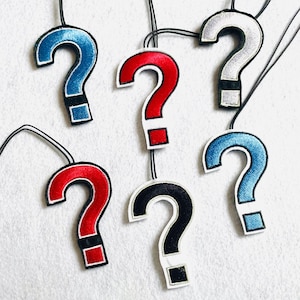 Könnte beinhalten: Eine Sammlung von Fragezeichen-Anhängern in verschiedenen Farben, darunter Rot, Blau, Silber und Schwarz. Jeder Anhänger hat eine schwarze Kordel zum Tragen. Die Fragezeichen sind mit einem weißen Rand bestickt.