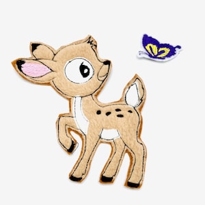 Könnte beinhalten: Ein rehfarbenes Filzreh mit schwarzen Punkten und einer weißen Brust, mit einem kleinen blauen und gelben Schmetterling. Das Reh hat schwarze Umrisse und rosa Innenohren. Der Schmetterling hat gelbe Details und einen weißen Umriss.