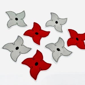 Könnte beinhalten: Sieben Ninja-Stern-Patches in Rot und Grau. Jeder Stern hat eine schwarze Mitte und einen weißen Rand. Die Patches sind auf einem weißen Hintergrund angeordnet. Geeignet zum Aufnähen auf Kleidung oder Accessoires.