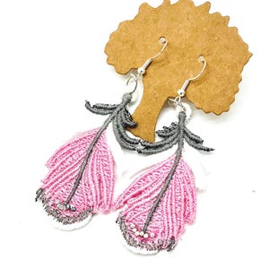 Könnte beinhalten: Ein Paar rosa-graue Federohrringe mit silbernen Haken. Die Ohrringe sind aus besticktem Stoff gefertigt und weisen ein detailliertes Federdesign auf. Die Federn haben einen rosa Körper, einen grauen Stiel und weiße und graue Akzente. Kleine Perlen sind unten angebracht.