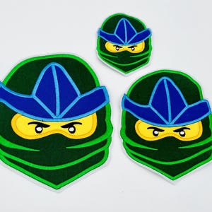 Könnte beinhalten: Drei gestickte Ninja-Gesichtspatches in verschiedenen Größen. Jeder Patch zeigt eine grüne Ninjamaske, eine blaue Kopfbedeckung und gelbe Augen. Die Patches sind grün umrandet und haben einen weißen Rand.