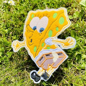Könnte beinhalten: Gestickter Aufnäher mit der Figur SpongeBob Schwammkopf in überraschter Pose. Der Aufnäher ist gelb mit grünen Punkten, blauen Augen und braunen Shorts. Die Ränder sind weiß.