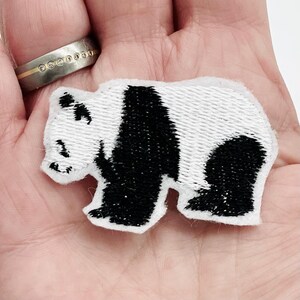 Könnte beinhalten: Gestickter Panda-Bären-Aufnäher mit schwarzen und weißen Details. Der Aufnäher zeigt ein Panda-Bären-Design mit weißem Hintergrund und schwarzen Akzenten. Ideal zum Aufnähen auf Kleidung, Taschen oder andere Stoffartikel.