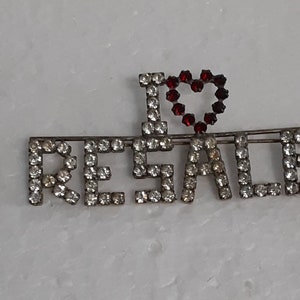 May include: A silver pin with rhinestones spelling out "I love resale" with a red rhinestone heart.