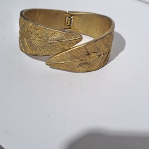 May include: A gold-toned metal bangle bracelet with a leaf design. The bracelet is hinged and opens to fit the wrist.
