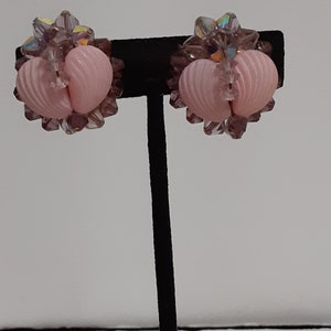 Może przedstawiać: Para zabytkowych kolczyków na klips. Każdy kolczyk ma jasno-różowy, sercowaty środek z żebrowaną fakturą, otoczony skupiskami małych, fasetowanych, fioletowych i iryzujących kryształków. Kolczyki są prezentowane na czarnym stojaku na białym tle.