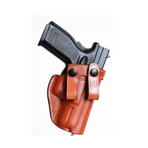 Könnte beinhalten: Ein braunes Lederholster mit einer schwarzen Pistole darin. Das Holster hat zwei Druckknöpfe an der Seite.