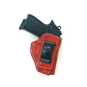 May include: A brown leather holster with a white stitching detail and a black clip. A black handgun is inside the holster.