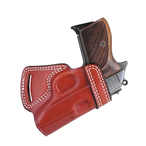 Könnte beinhalten: Ein rotes Lederholster mit weißen Nähten für eine Pistole mit einem Holzschaft. Die Pistole hat eine silberne Oberfläche und die Worte "TURK" sind an der Seite sichtbar.