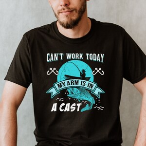 May include: A black t-shirt with a blue graphic of a fisherman in a boat with a fish jumping out of the water. The text on the shirt reads "Can't work today, my arm is in a cast".