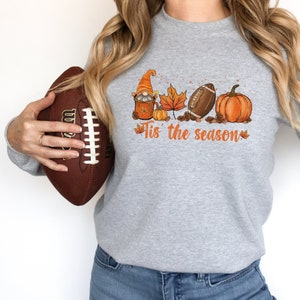 Op de afbeelding: Een lichtgrijze sweatshirt met een herfstthema. Het ontwerp bevat een kabouter, een esdoornblad, een American football en een pompoen. De tekst "Tis the season" is in oranje geschreven. Een bruine American football wordt vastgehouden.