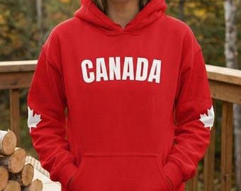 May include: A red hooded sweatshirt with the word "CANADA" in white block letters across the chest. The sweatshirt has a kangaroo pocket and a white maple leaf on the left sleeve. A person is wearing the sweatshirt, standing outside with a dog.