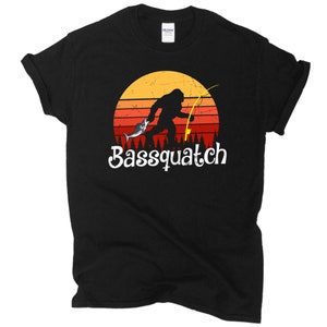 May include: Black t-shirt with a retro sunset graphic featuring a Bigfoot holding a fish and a fishing rod. The text "Bassquatch" is printed below the graphic.
