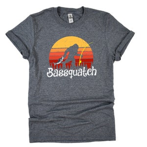 May include: A dark heather grey t-shirt with a retro-style graphic of a Bigfoot holding a fishing rod and reeling in a fish. The graphic is set against a sunset background with red, orange, and yellow stripes. The text "Bassquatch" is printed in white below the graphic.
