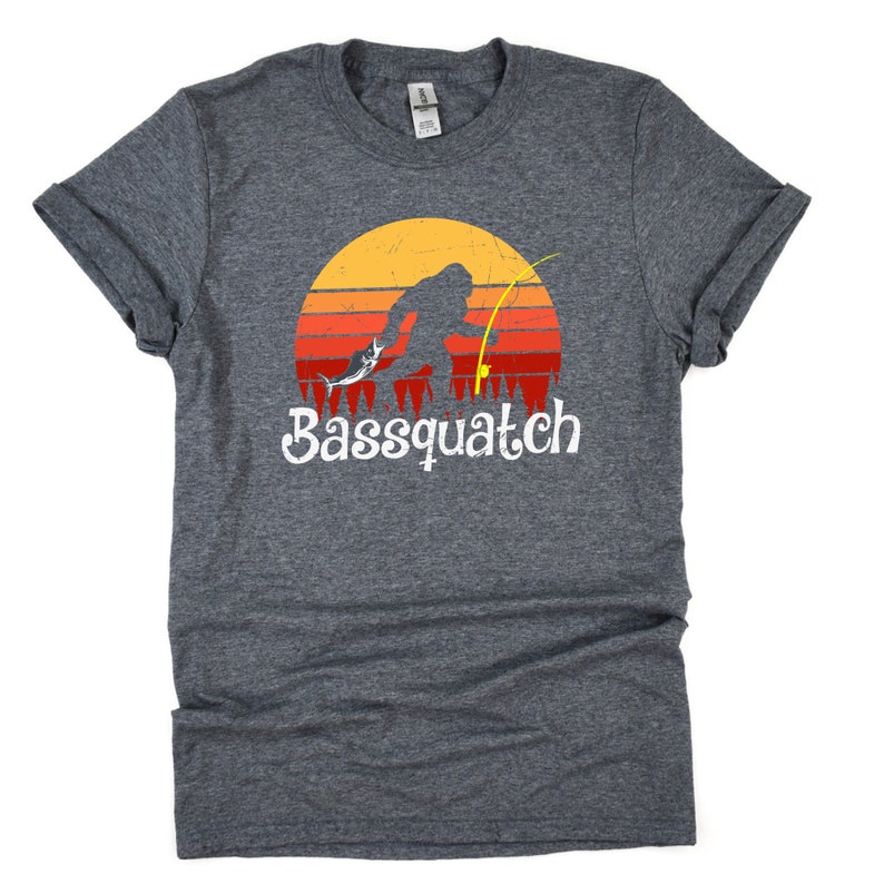 May include: A dark heather grey t-shirt with a retro-style graphic of a Bigfoot holding a fishing rod and reeling in a fish. The graphic is set against a sunset background with red, orange, and yellow stripes. The text "Bassquatch" is printed in white below the graphic.