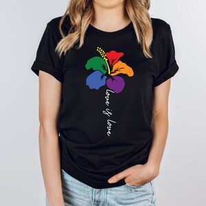 Könnte beinhalten: Schwarzes T-Shirt mit einer Regenbogen-Hibiskus-Blume und dem Aufdruck "love is love" auf der Vorderseite.