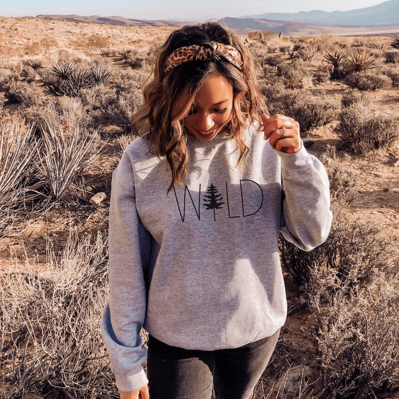 May include: A gray sweatshirt with the word "WILD" printed on the front in black with a small pine tree graphic in the middle of the word. The model is wearing a leopard print headband.