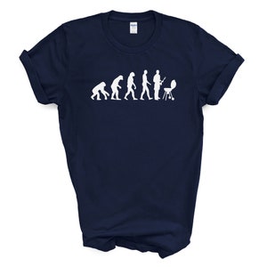 May include: A navy blue t-shirt with a white graphic of the evolution of man, ending with a man grilling. The graphic shows a series of stick figures, starting with a primitive ape and ending with a man standing next to a grill.