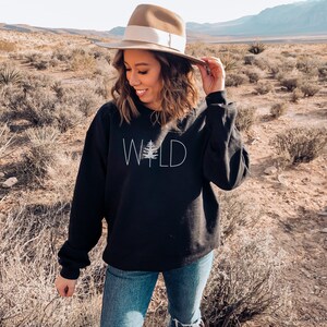 May include: A black sweatshirt with the word "WILD" printed on the front in white with a stylized pine tree replacing the "I". The model is wearing a brown fedora and blue jeans.