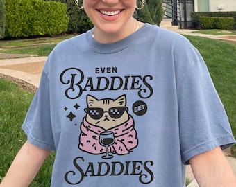 May include: A blue short-sleeve t-shirt featuring a graphic of a cat wearing sunglasses and wrapped in a pink blanket while holding a glass of wine. The text reads "EVEN BADDIES GET SADDIES" in black lettering. The shirt has a crew neckline.