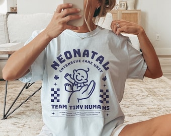 May include: A light blue short-sleeve t-shirt featuring a dark blue graphic print that reads NEONATAL INTENSIVE CARE UNIT TEAM TINY HUMANS with an illustration of a baby being held in a hand, accompanied by white shorts and white socks.