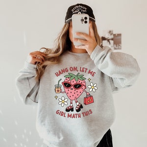 May include: Gray sweatshirt with a cartoon strawberry wearing sunglasses and holding a shopping bag. The text on the sweatshirt reads "Hang on, let me girl math this."