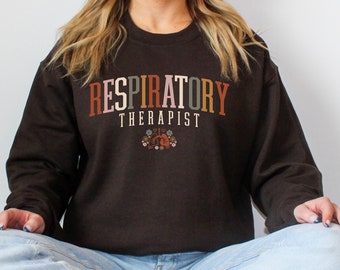 May include: Dark chocolate-colored sweatshirt with the text "RESPIRATORY THERAPIST" in a retro font. The design includes a floral graphic. The sweatshirt is a crew neck style.