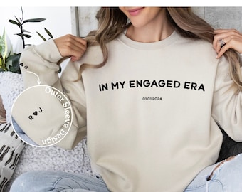 May include: Cream-colored sweatshirt with the text "IN MY ENGAGED ERA" and the date 01.01.2024 printed in black. The outer sleeve design features initials "R ♥ J". The sweatshirt is a casual style.